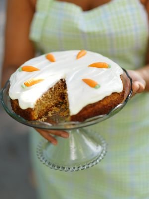 Morotskaka med vit frosting och morotsdekoration, serverad på glasfat hållet av person i rutig förkläde.