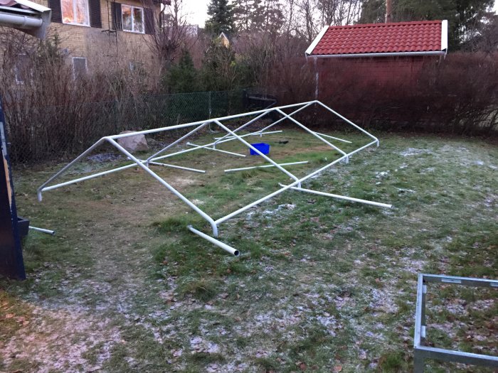 Nedmonterad snickarboa av metallrör på en frostig gräsmatta med hus och buskar i bakgrunden.