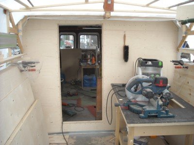 Utsikt från ett båtdäck under renovering med träväggar och en arbetsbänk med verktyg, leder in till ett annat utrymme.