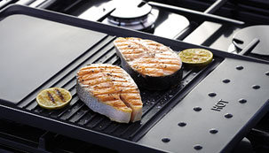Induktionshäll med teppanyaki stekbord och laxfiléer på grillen, omgiven av gasbrännare.