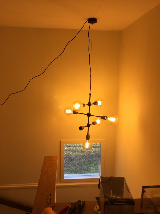 Hängande taklampa med flera glödlampor i ett rum under renovering med synligt fönster och verktyg.