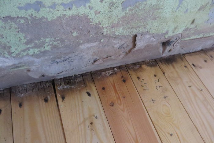 Skador på nedre delen av en innervägg med lossnande tapet och puts samt mörknade trägolvbrädor längst in vid väggen.
