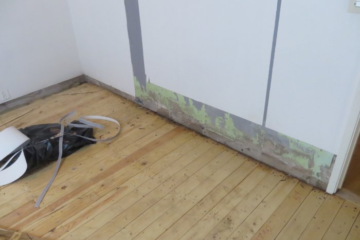 Skadad vägg med lossnad tapet och plywood samt mörknade golvbrädor i ett hörn av rummet.