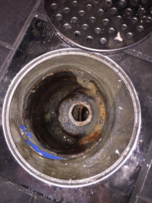 Sliten golvbrunn med rost och avlagringar i en nyköpt lägenhet, omringad av mörka klinkerplattor.