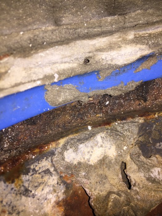 Närbild på rostig järnbalk med fuktskador och blå plastledning.
