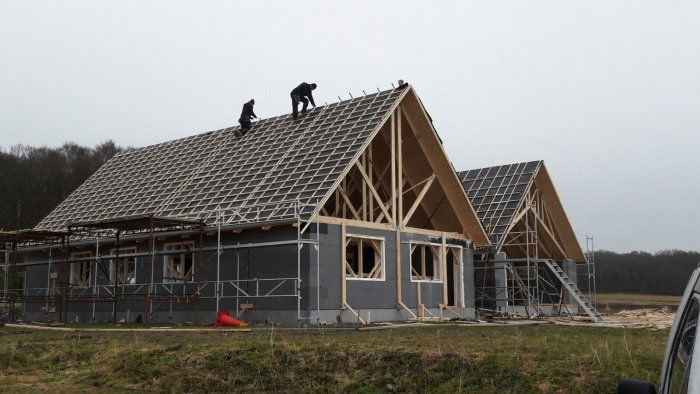 Två arbetare installerar takstolar på ett hus under konstruktion i dagsljus.