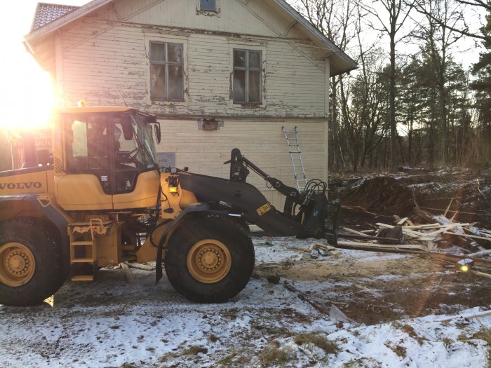 En Volvo hjullastare framför ett slitet hus med lite snö och röjningsarbete synligt i solljus.