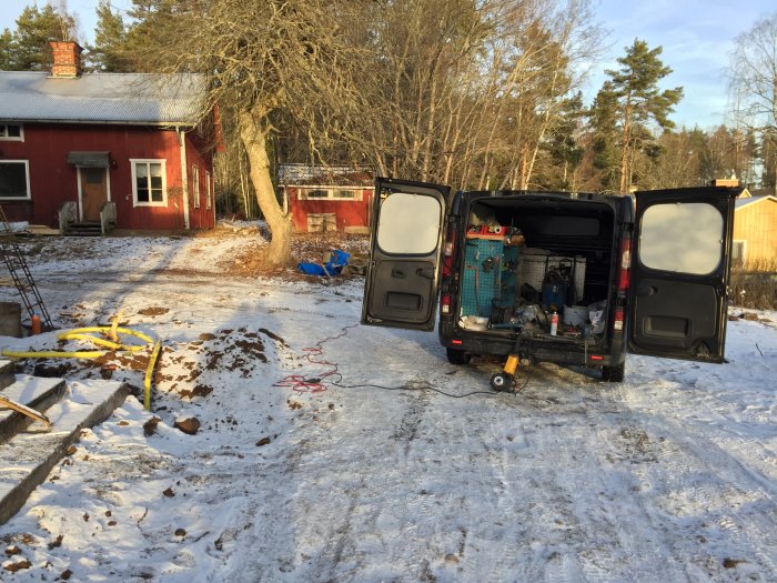 Byggarbetsplats med öppen verktygsfylld skåpbil framför röd stuga och byggmaterial i vintersol.