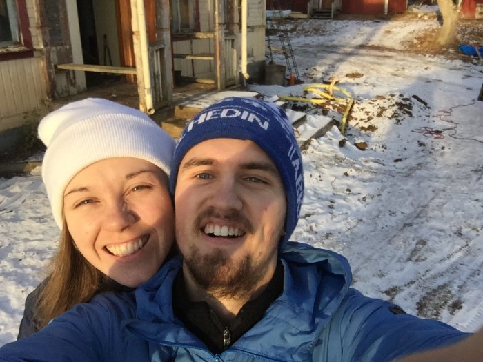 Två leende personer tar en selfie framför ett hus under renovering med verktyg och material synliga.