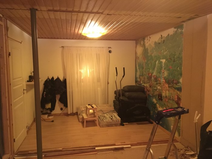 Rum under renovering med träningscykel, takpanel och väggmålning med djurmotiv.