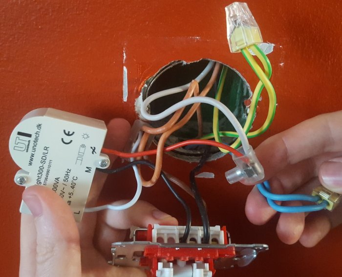 Händer som håller en elektrisk dimmerkoppling framför en öppen väggdosa med synliga ledningar.