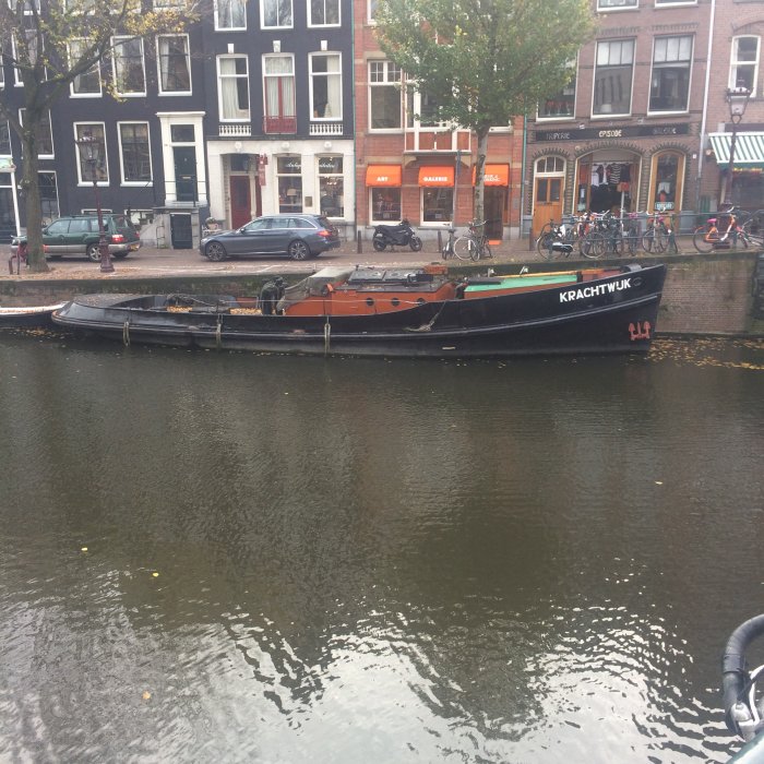 En äldre båt med namnet "KRACHTWIJK" förtöjd vid en kanal, med stadsmiljö i bakgrunden.