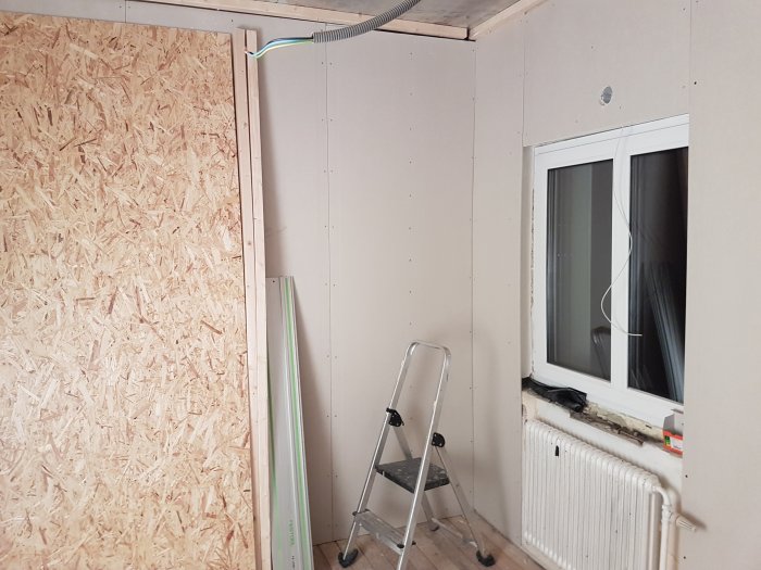 Omfattande renovering av ett lekrum med oskyddat fönster, osammanhängande väggpaneler och öppen golvyta för golvvärme.