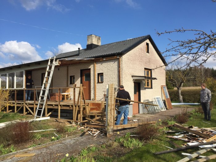 Renovering av en gammal altan på ett hus, med tre personer som arbetar och diverse byggmaterial utspridda på marken.