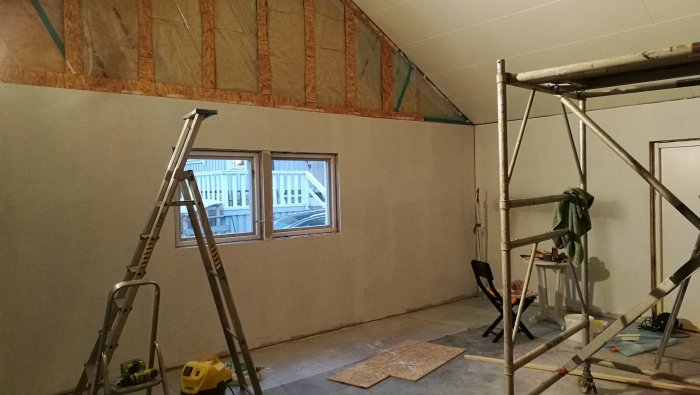 Renoveringsarbete i rum med målat halvtak och grundmålade väggar, stege och byggställning synliga.