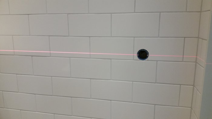 Vägg med vita kakelplattor och en svart röranslutning, med en rosa laserlinje för utmätning och positionering.