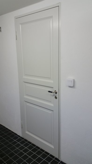 Nyinstallerad vit dörr med handtag, lås och ljusströmbrytare på en vit vägg ovanför svartvitt klinkergolv.