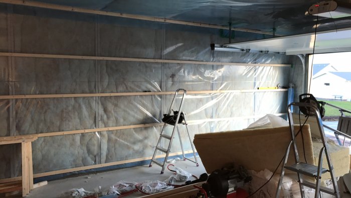 Garage under renovering med isolering och plast uppsatt på väggar, reglar, och rörigt byggmaterial i förgrunden.