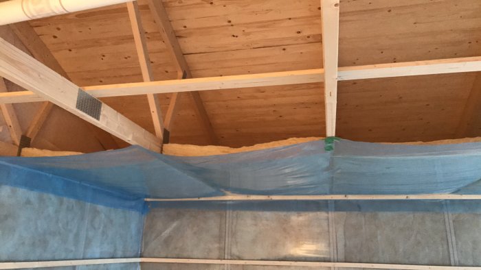 Monteringsprocess i ett garage med blå plastfolie och isolering i taket, mellan träreglar och takpaneler.
