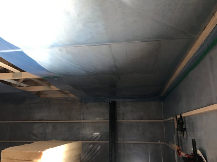 Plastfolie som hänger i taket i ett garage under ombyggnation, med träreglar och isoleringsmaterial synligt.