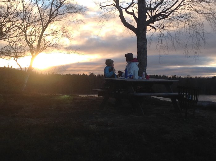 Två personer och ett barn vid ett picknickbord nära en sjö under en solnedgång.