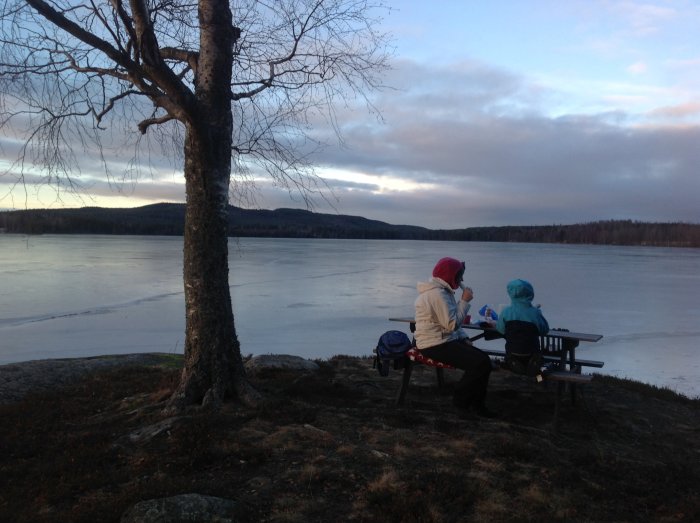 Två personer fikar på en bänk vid en frusen sjö med träd och målningsfull himmel i bakgrunden.
