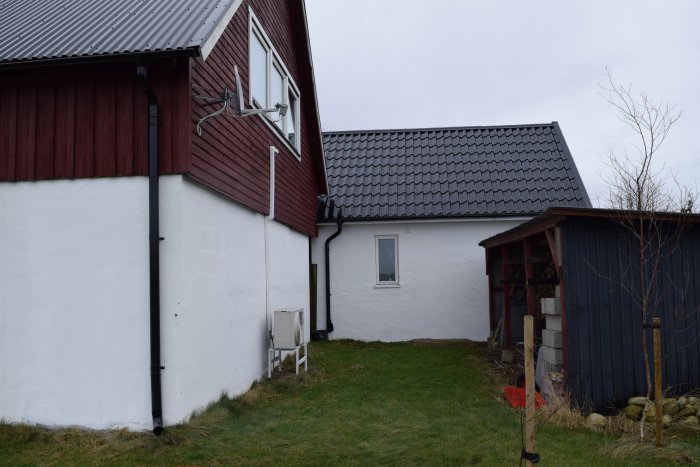 Punainen talo valkoisella kivijalalla, vierekkäin pienempi rakennus, musta seinä. Edessä nurmikkoa.