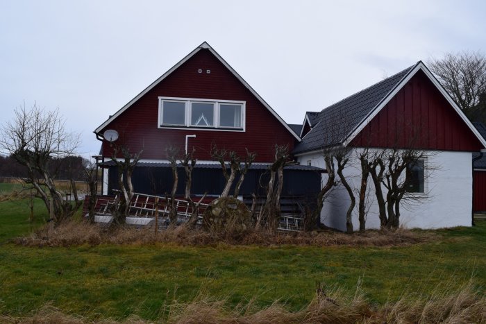 Punainen talo, peltikatos ja karsittuja puita edessä. Pihalla rakennustikkaat ja ruohonleikkuri. Taustalla pelto, kuvattu pilvisenä päivänä.