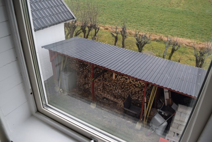 Näkymä ikkunasta, joka näyttää peltikaton alla olevan puupinon vieressä pihalla olevia työkaluja ja roskakori. Kuvattu ylhäältäpäin.