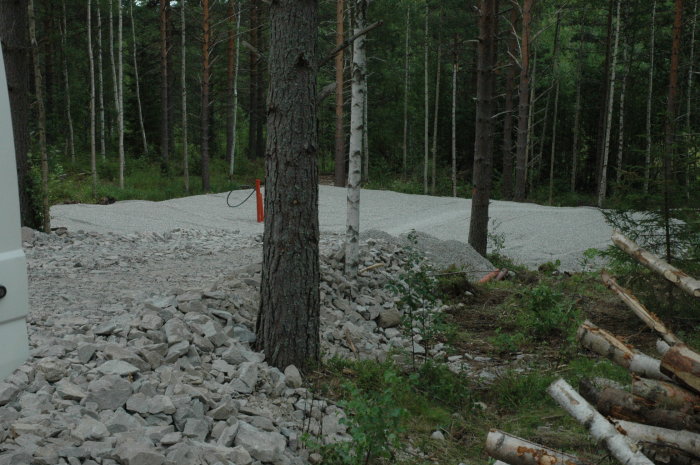 Tomt med påbörjat markarbete och högar av grus och sten, omgiven av skog.