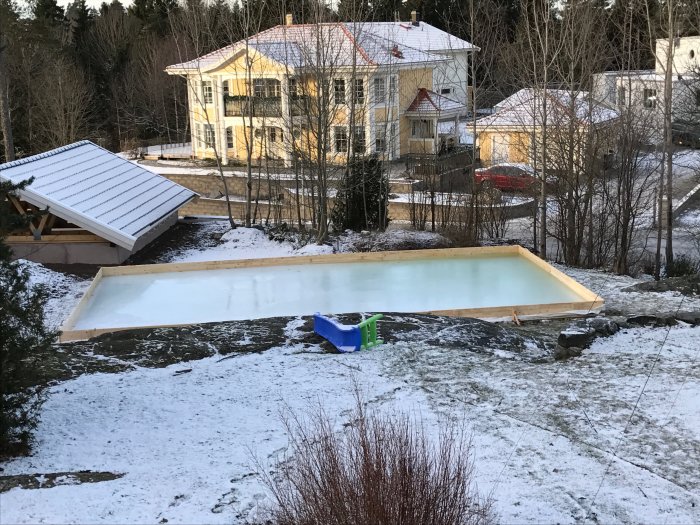 Hemmagjord isbana med träram i en snöig trädgård, nära gula hus och träd.