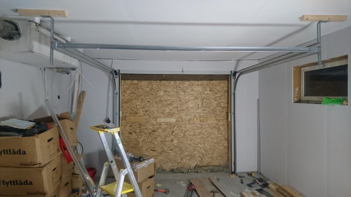 Nyinstallerad garageport med öppningsmekanism framför en tillfällig OSB-vägg i ett rörigt garage.