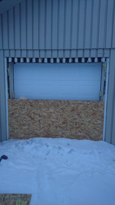 Ny monterad garageport ovanför OSB-skiva i en byggnad med grå panel och snö framför.