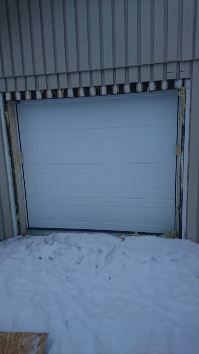 Nyinstallerad vit garageport i en byggnad med träfasad, omgiven av snö och ofärdiga kanter.