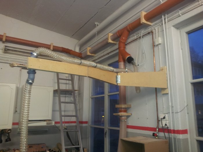 Inomhusvy av rörledningar och provisorisk trästödning vid en byggplats.