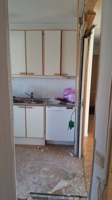 Kök under renovering med öppna skåputrymmen och byggbråte på golvet, sett genom delvis rivet väggparti.