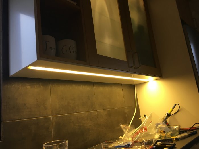 Köksskåp med LED-belysning i frästa spår och opalvit plexiglasöverdrag, verktyg och material på arbetsyta.