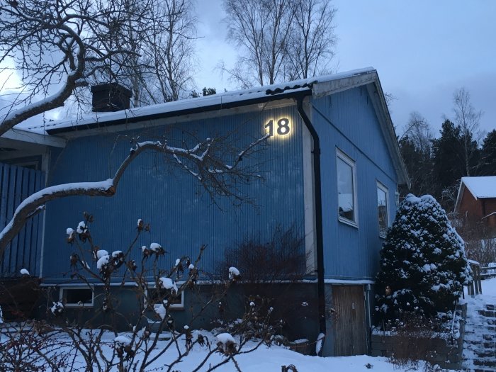 Illuminerade husnummer '18' på en blå träfasad med snötäckt landskap i skymningen.