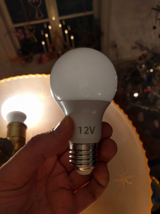 Hand håller en LED-lampa märkt med 12V framför en suddig bakgrund med juldekorationer.