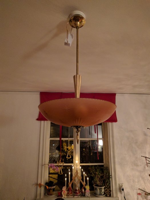 Retro taklampa med gulddetaljer och en stor skärm, hängandes i ett rum med juldekorationer i fönstret.