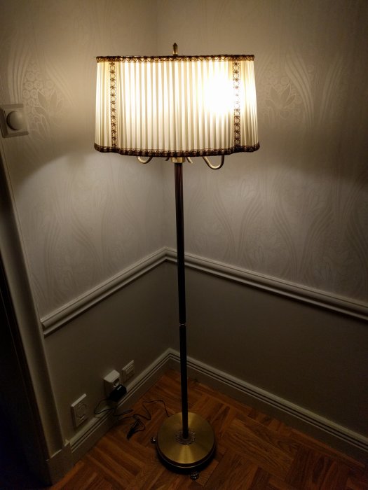 Golvlampa tänd i ett hörn med ornament och tapet, vid vägguttag på parkettgolv.