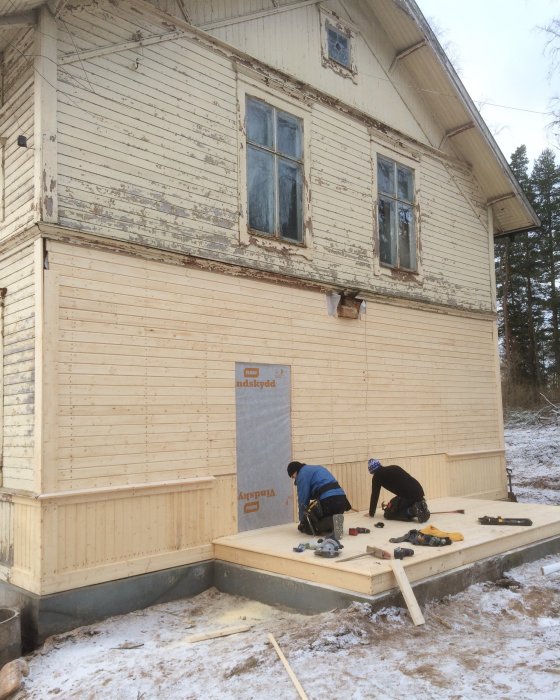 Två personer monterar nytt verandagolv på gammalt trähus, verktyg utspridda, en del panel återstår att montera.