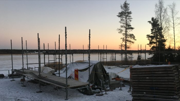 Byggnadsställningar vid solnedgång vid en frusen sjö med träd i bakgrunden.