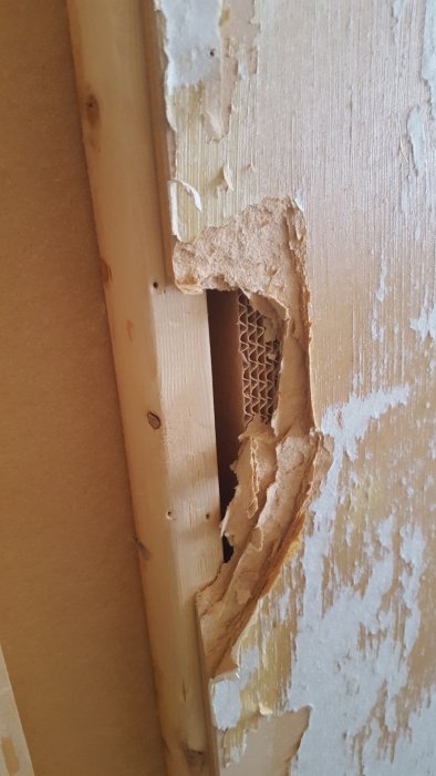 Innervägg med skada som avslöjar veckad papp mellan vertikala träreglar och MDF-panel.