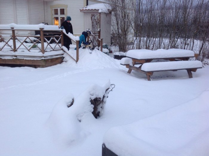 Två personer skottar snö i en trädgård med snötäckta utemöbler.