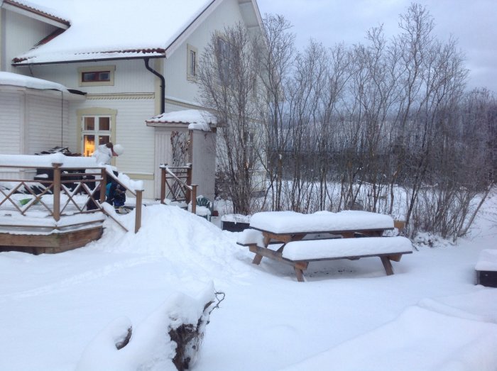 Snötäckt trädgård med person som skottar snö nära ett hus och picknickbord under vinterväder.