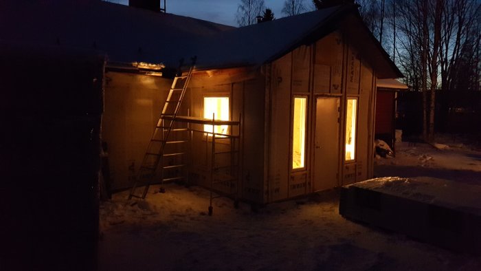 Byggplats i skymningen med upplysta fönster och en temporär dörr på ett hus under konstruktion.