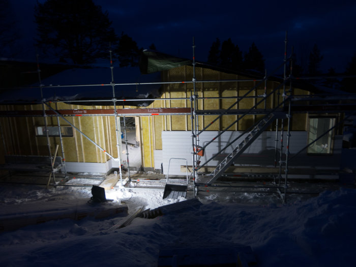 Byggarbetsplats vid skymning med uppspikad panel och byggnadsställningar runt ett hus i vintersnö.