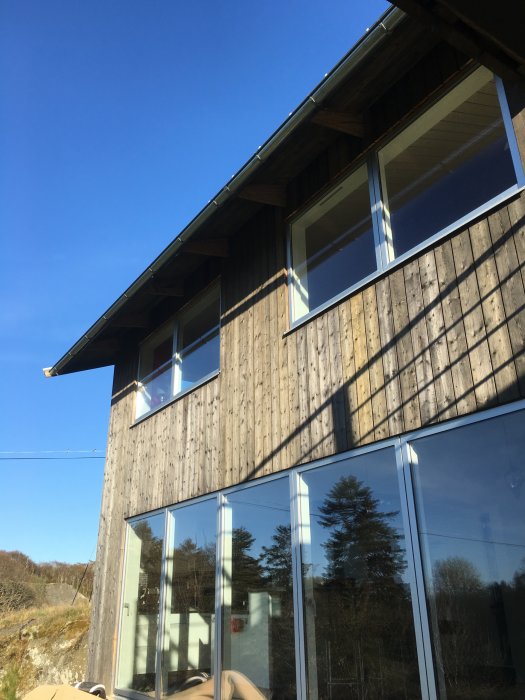 Husfasad med väderbiten träpanel och stora fönsterpartier under en klarblå himmel.