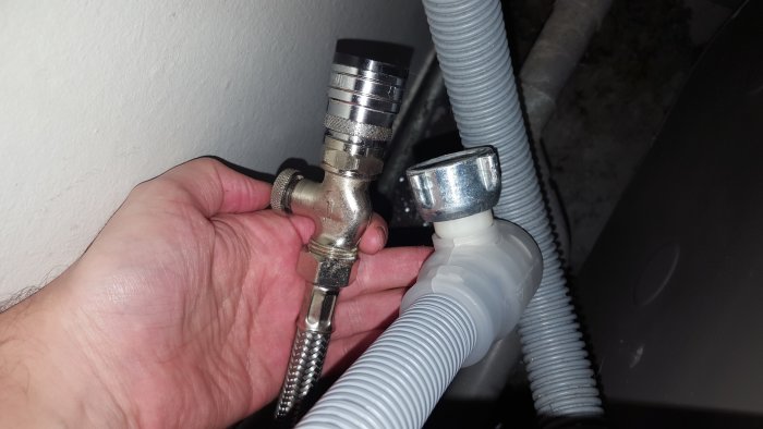 En hand håller en tvättmaskinslang nära en okopplad metallventil och en ansluten vit plastslang.